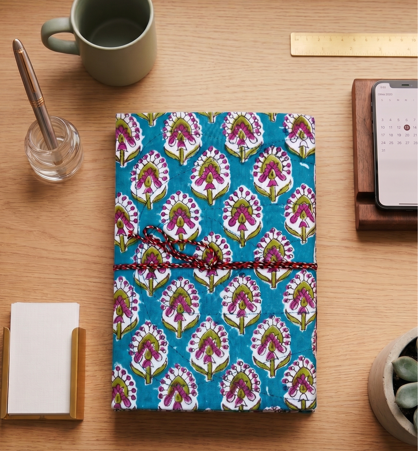 A closed notebook with a vibrant blue, pink, and green floral block print pattern, tied with a red cord, displayed on a wooden desk surrounded by office props like a mug, phone, and notepad.
