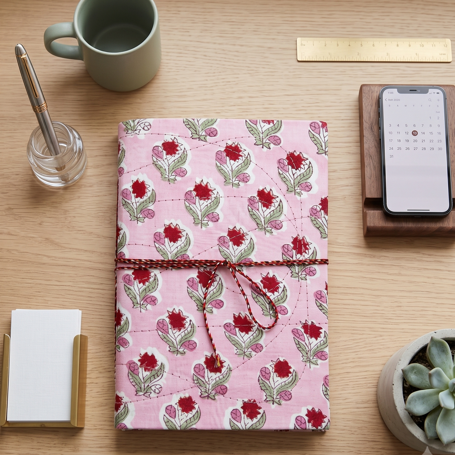 A pink floral notebook or planner cover with a red string tie closure, displayed on a light wood desk alongside a mug, phone on a stand, card holder, and small plant.