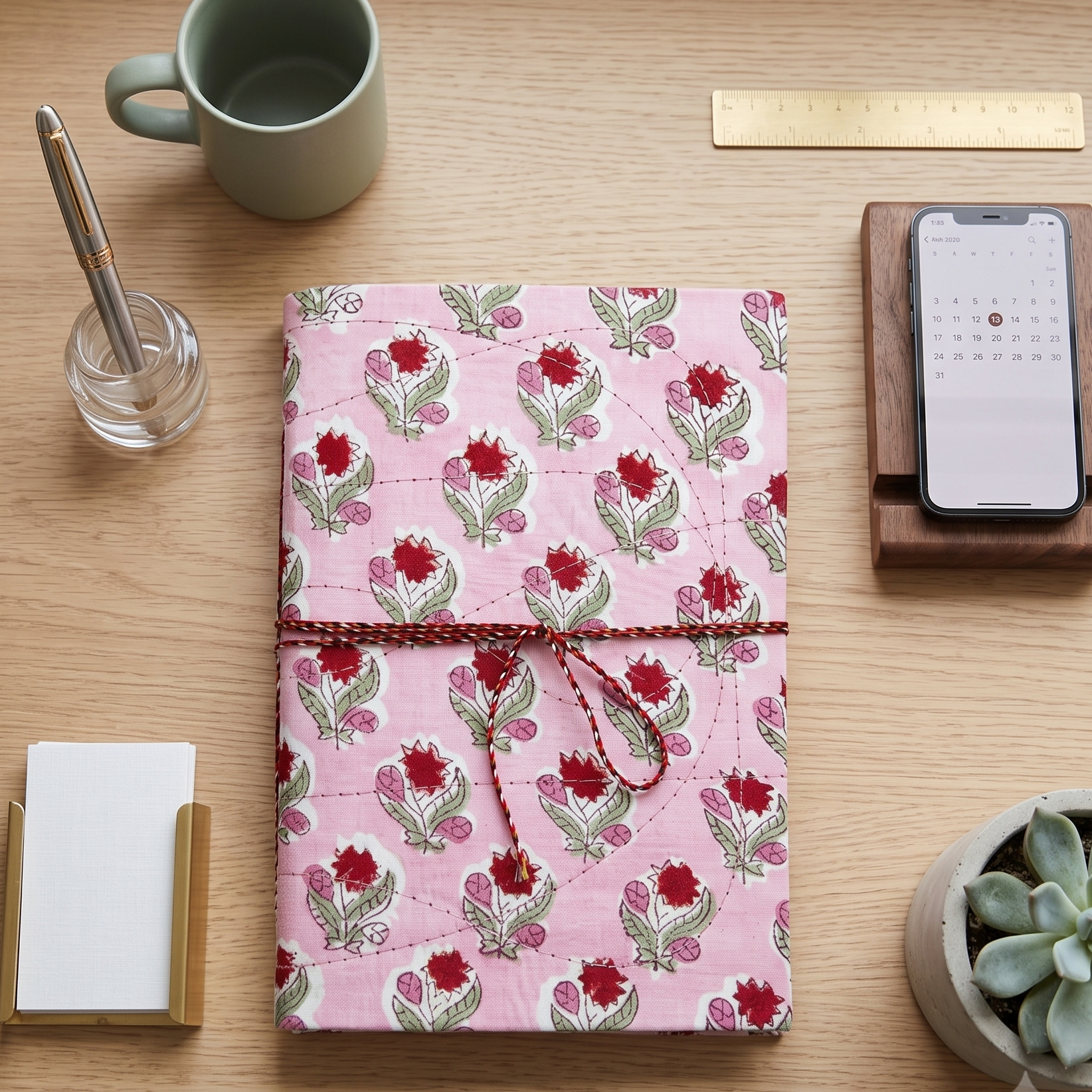 A pink floral notebook or planner cover with a red string tie closure, displayed on a light wood desk alongside a mug, phone on a stand, card holder, and small plant.