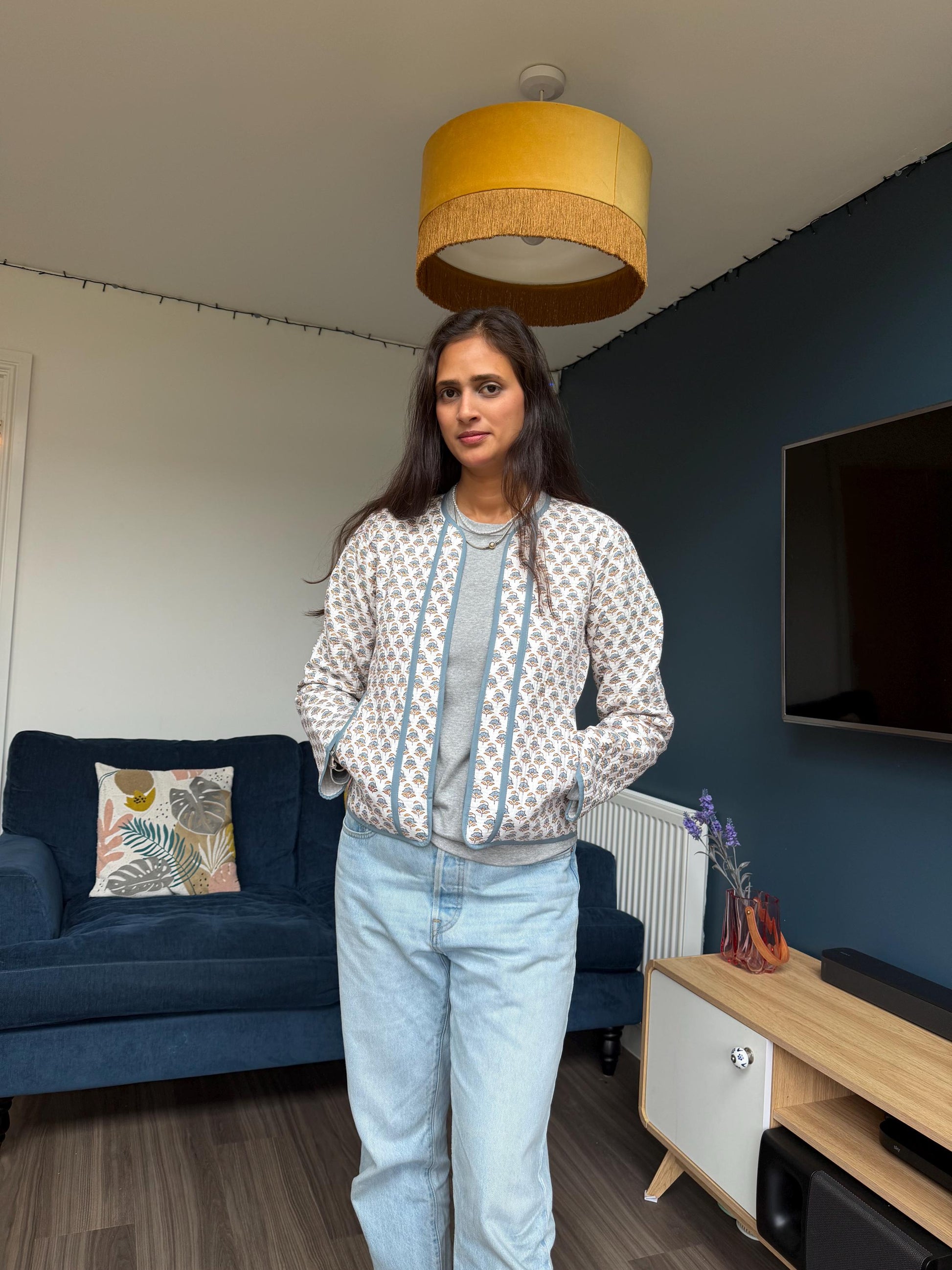 May include: A patterned jacket with a light blue trim and a gray shirt. The jacket has a white background with a repeating blue design. The person is wearing light blue jeans. The background includes a blue sofa and a wooden cabinet.
