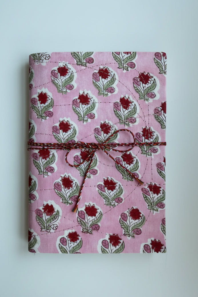 A pink fabric-covered notebook or journal with a repeating block-print floral pattern in red, green, and white, tied with a brown twine wrap.