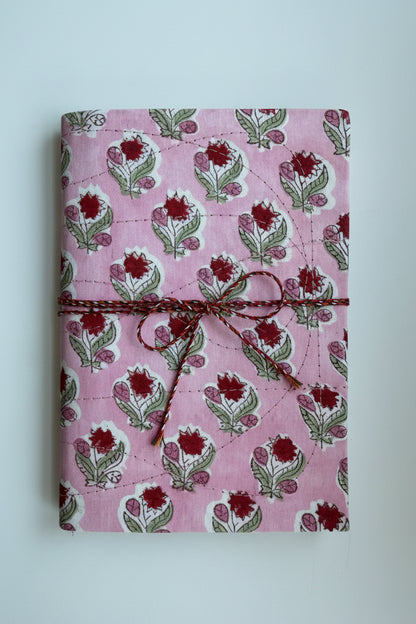 A pink fabric-covered notebook or journal with a repeating block-print floral pattern in red, green, and white, tied with a brown twine wrap.